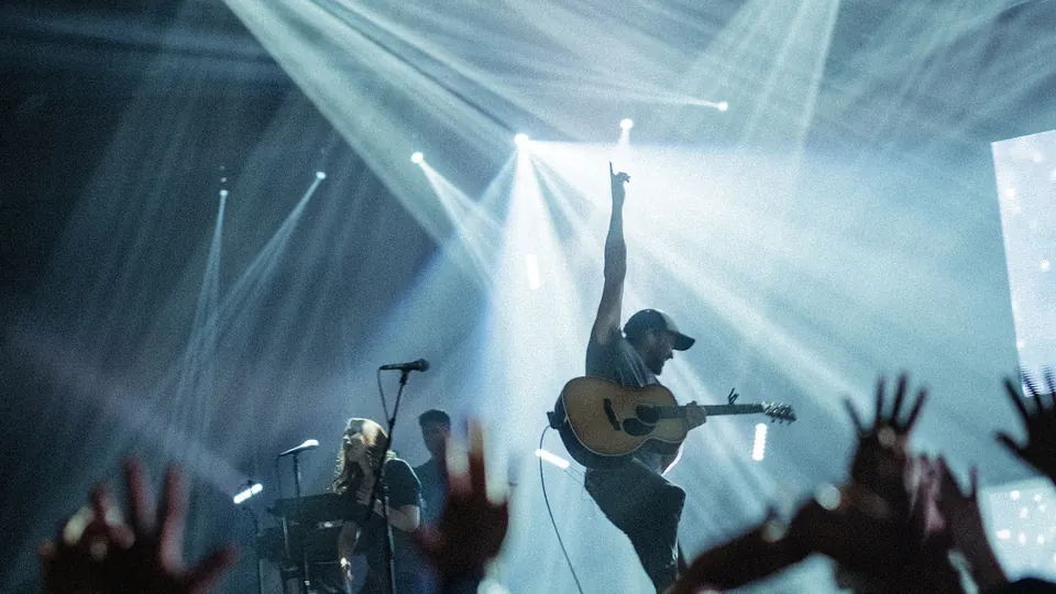 Worship leader with guitar on stage under bright beams of light